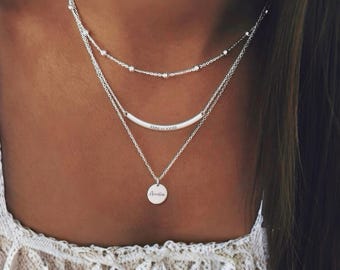 Op de afbeelding: Een zilveren gelaagde ketting set. De bovenste laag heeft kleine kralen, de middelste laag een gebogen staaf met tekst en de onderste laag een ronde hanger met de naam "Amelia".