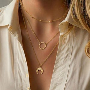 Op de afbeelding: Een set gouden kettingen die over elkaar worden gedragen. De bovenste ketting heeft kleine kralen, de middelste een eenvoudige cirkel en de onderste een halve maan hanger. De kettingen worden gedragen over een wit overhemd.