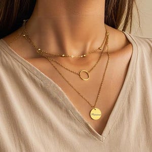 Könnte beinhalten: Ein Satz goldener Halsketten in mehreren Schichten. Die oberste Kette hat kleine Perlen, die mittlere einen einfachen Kreisanhänger und die untere einen runden Anhänger mit eingraviertem Text. Präsentiert auf einem beigefarbenen ärmellosen Oberteil.