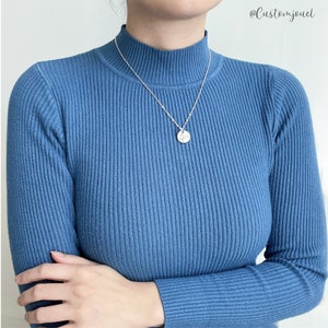 May include: A silver-toned necklace featuring a small, circular pendant with a delicate line drawing of a figure. The necklace is worn over a blue, long-sleeved, ribbed mock-neck sweater. The text @Customjouel is visible in the upper right corner.