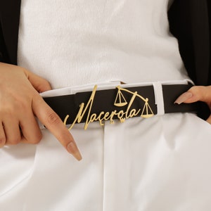 Puede incluir: Un cinturón de cuero negro con una hebilla de metal dorado con las palabras "Macerola Law" y un diseño estilizado de balanza de la justicia.