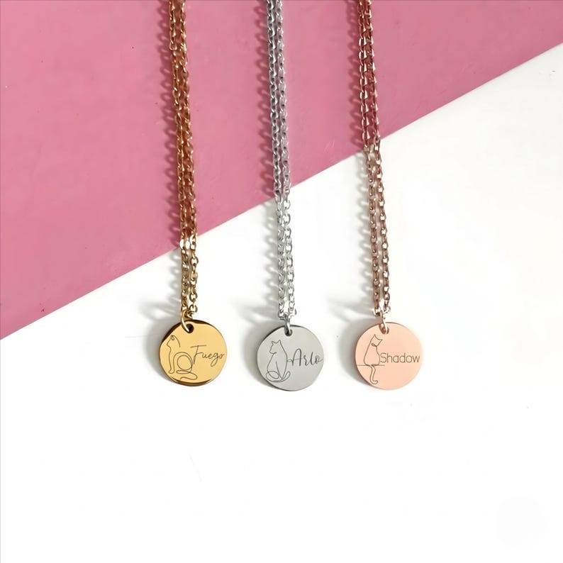 May include: Three necklaces with round metal pendants hanging from delicate chains. The left pendant is gold with a cat outline and the name Fuego. The center pendant is silver with a cat outline and the name Arlo. The right pendant is rose gold with a cat outline and the name Shadow.