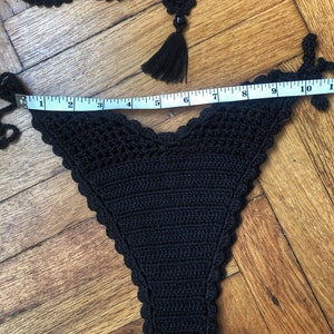 May include: Black crocheted bikini bottom with a scalloped edge. The bottom is measured with a tape measure, showing a length of 10 inches.
