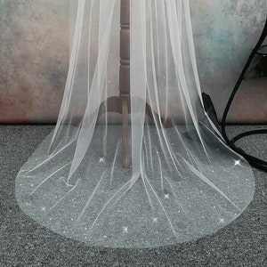 May include: A delicate white bridal veil with shimmering details. The veil is displayed on a wooden dress form, highlighting its flowing form. The lower edge of the veil features a concentration of sparkling embellishments.