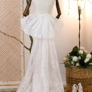 Peut inclure: Un voile de mariée en dentelle blanche avec un grand nœud à l'arrière. Le voile est drapé sur un mannequin blanc et est présenté sur un tapis tissé brun clair.