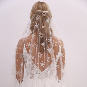 Può includere: Un velo da sposa bianco con un motivo a fiocco di neve. Il velo è in tulle ed è decorato con delicati ricami a fiocco di neve.