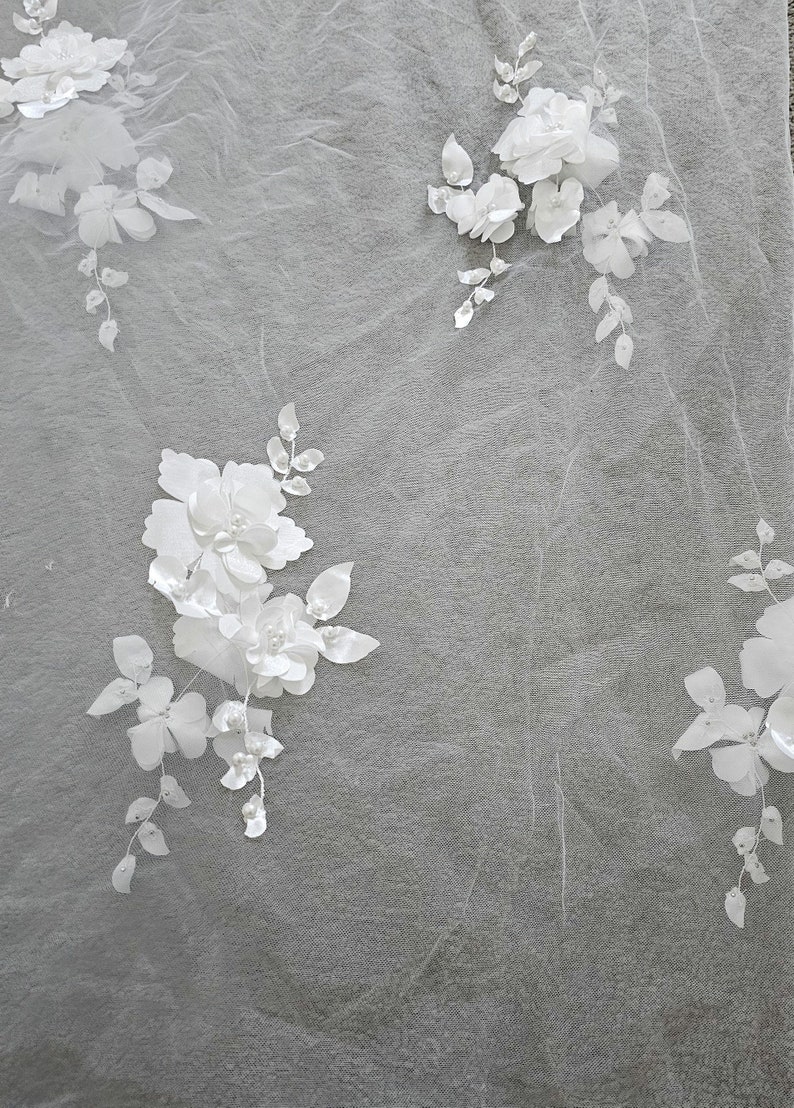 May include: White floral lace fabric with delicate 3D flowers and pearl accents. The fabric is sheer and lightweight, perfect for adding a touch of elegance to any project.