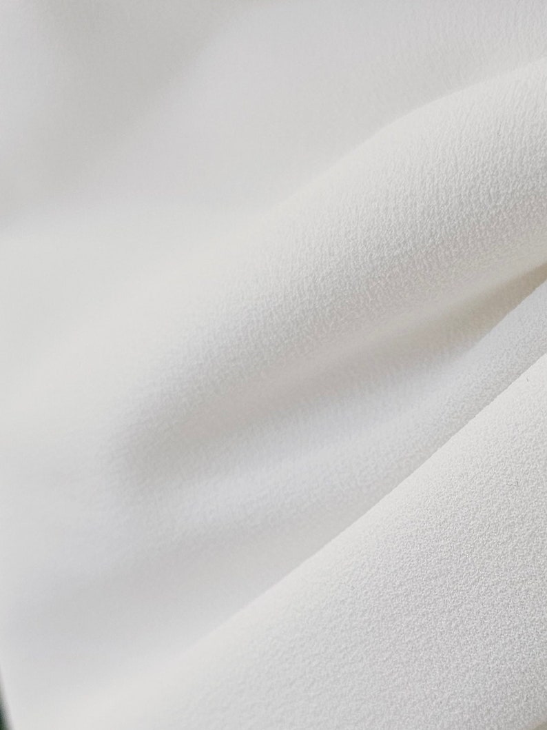 May include: Close-up of a white fabric with a textured surface. The fabric is soft and smooth, with a subtle sheen.