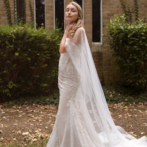 May include: A bride wearing a white wedding dress with a long veil. The dress has a fitted bodice and a flowing skirt. The bride is standing in front of a brick building with windows.