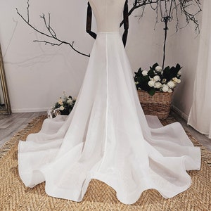 Puede incluir: Un vestido de novia de tul blanco con una falda fluida. El vestido está en un maniquí y se muestra en una alfombra marrón.