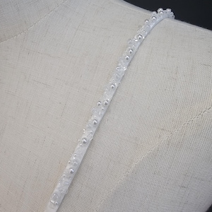May include: A white, beaded strap with small, round, pearl-like embellishments. The strap is pinned to a mannequin with a yellow pin. The background is a neutral color.