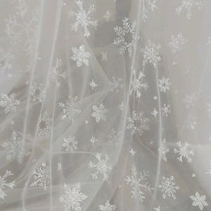 May include: White tulle fabric with a snowflake pattern. The snowflakes are made of glitter and are scattered randomly across the fabric.