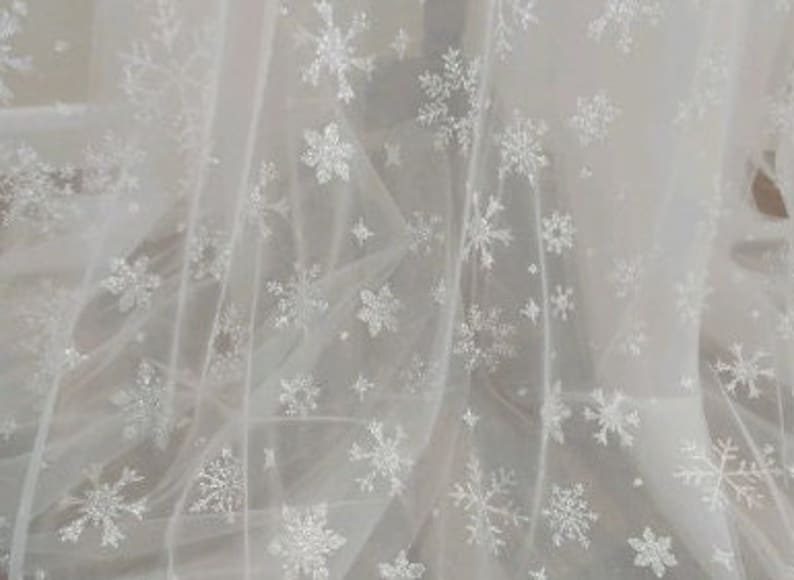 May include: White tulle fabric with a snowflake pattern. The snowflakes are made of glitter and are scattered randomly across the fabric.