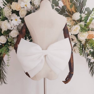 May include: A large white bow made of fabric, tied around a white mannequin. The bow is positioned on the back of the mannequin, with the ends of the bow hanging down.