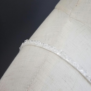 May include: A close-up of a white, beaded headband. The headband is curved and sits on a white fabric surface. The beads are clear and small, adding a delicate touch to the accessory.