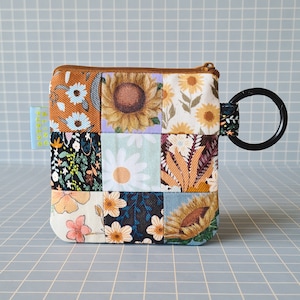 Op de afbeelding: Een klein patchwork stoffen ritsvakje met een bloemen- en zonnebloemprint. Het vakje heeft een bruine rits en een zwarte metalen ring die aan de rits bevestigd is. Het vakje ligt op een grijze ruitjesachtergrond.