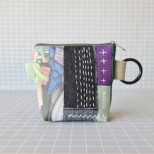 Può includere: Una piccola pochette con cerniera in tessuto patchwork con un pannello in tessuto bianco e nero, un pannello in tessuto viola con segni più bianchi e un pannello in tessuto verde con testo bianco. La pochette ha un anello di metallo nero attaccato a una cinghia in tessuto verde.