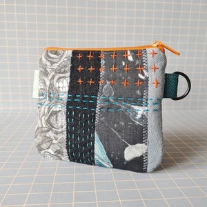 Peut inclure: Une petite pochette zippée en patchwork avec un imprimé de crâne noir et blanc, des coutures bleues et orange et une sangle verte avec un anneau métallique.