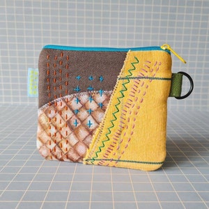 Peut inclure: Une petite pochette zippée en patchwork jaune, marron et blanc avec un tirette de fermeture éclair verte et un anneau métallique noir. La pochette est décorée de coutures colorées.