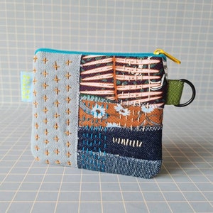 Peut inclure: Une petite pochette zippée en patchwork avec un motif en tissu bleu, marron et blanc. La pochette a une fermeture éclair dorée et une sangle verte avec un anneau argenté.