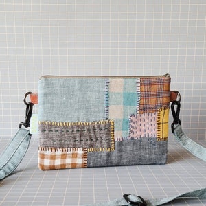 Puede incluir: Un bolso bandolera de tela patchwork con una correa azul y blanca. El bolso está hecho de telas de diferentes colores, incluyendo azul, marrón, blanco y rosa.