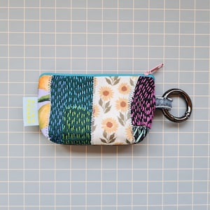 Può includere: Una piccola pochette con cerniera in tessuto patchwork con stampa di girasole, stampa di limone e stampa geometrica verde e blu. La pochette ha un anello di metallo nero attaccato al tiretto della cerniera.