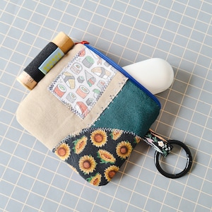 Peut inclure: Une petite pochette multi-color patchwork avec une fermeture éclair et un anneau noir attaché. La pochette présente une variété de motifs, notamment des sushis, des tournesols et un tissu vert uni. La pochette est posée sur une surface grise quadrillée.