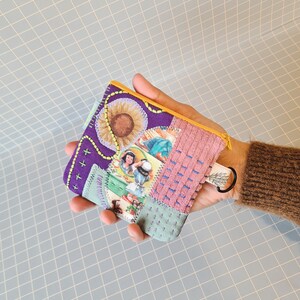 Peut inclure: Une petite pochette en tissu violet avec une fermeture éclair et un anneau métallique. La pochette est décorée d'un motif patchwork avec une variété de couleurs et de motifs, y compris un motif floral, une fille en robe et un tournesol.
