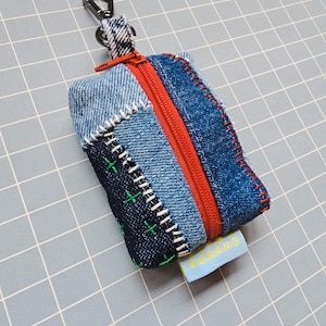 Op de afbeelding: Een kleine, rechthoekige denim pouch met een rode rits en een zwarte metalen karabijnhaak. De pouch is gemaakt van patchwork denim in verschillende blauwtinten, met witte en groene stiksels. Een klein label met "PURANG" is bevestigd.