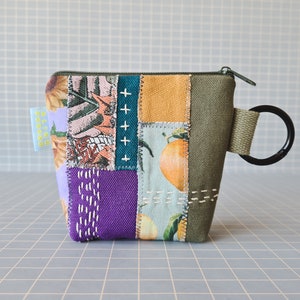 Peut inclure: Une petite pochette zippée en tissu patchwork avec une base en toile verte et une poignée en anneau. La pochette est décorée de tissus colorés variés, dont du violet, du rose, du jaune et du vert. La pochette se ferme par une fermeture éclair et porte une petite étiquette sur laquelle est inscrit "Building Blocks".
