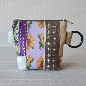Peut inclure: Une petite pochette zippée en patchwork avec un motif en tissu violet, vert et marron. La pochette est ornée d'un imprimé de tournesols et d'un petit anneau attaché au tirette de la fermeture éclair.