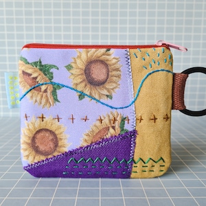 Peut inclure: Une petite pochette zippée en patchwork de couleur violette, jaune et marron avec un anneau noir attaché. La pochette a un motif floral avec des tournesols et est décorée de coutures.
