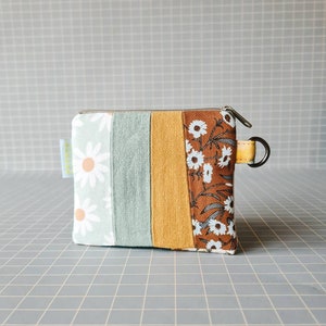 Peut inclure: Une petite pochette rectangulaire zippée avec un extérieur en tissu rayé brun, vert et jaune. La pochette a un imprimé de marguerites blanches d'un côté et un imprimé floral brun de l'autre. La pochette a un anneau métallique attaché à la fermeture éclair.