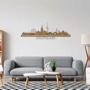 Op de afbeelding: Een houten wandkunstwerk dat de skyline van Stuttgart afbeeldt. De skyline is gemaakt van hout en toont de meest iconische gebouwen van de stad, waaronder de Fernsehturm, het Neues Schloss en de Staatsgalerie. Het kunstwerk is gemonteerd op een witte muur boven een grijze bank en een witte salontafel. Het woord "STUTTGART" staat in grijs onder de skyline.