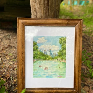 K&ouml;nnte beinhalten: Ein gerahmtes Aquarellbild, das ein t&uuml;rkisfarbenes Schwimmbecken umgeben von gr&uuml;nen B&auml;umen und einer Skyline unter einem blauen Himmel mit wei&szlig;en Wolken zeigt. Menschen schwimmen im Wasser. Die K&uuml;nstlersignatur Cochran ist in Pink.