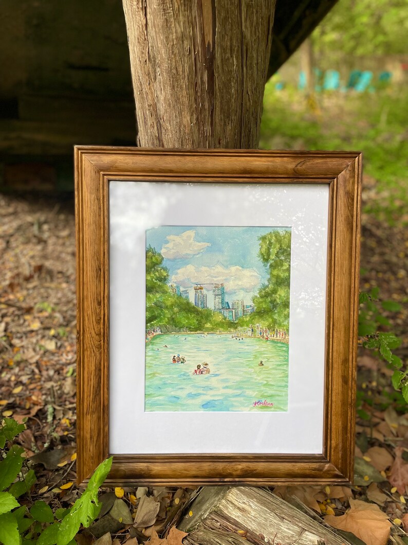 K&ouml;nnte beinhalten: Ein gerahmtes Aquarellbild, das ein t&uuml;rkisfarbenes Schwimmbecken umgeben von gr&uuml;nen B&auml;umen und einer Skyline unter einem blauen Himmel mit wei&szlig;en Wolken zeigt. Menschen schwimmen im Wasser. Die K&uuml;nstlersignatur Cochran ist in Pink.