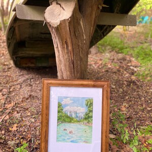K&ouml;nnte beinhalten: Ein gerahmtes Aquarellbild, das ein leuchtend blaues Schwimmbecken zeigt, umgeben von &uuml;ppigen gr&uuml;nen B&auml;umen und einer fernen Stadtsilhouette unter einem bew&ouml;lkten Himmel. Das Kunstwerk befindet sich in einem Holzrahmen mit wei&szlig;em Passepartout.