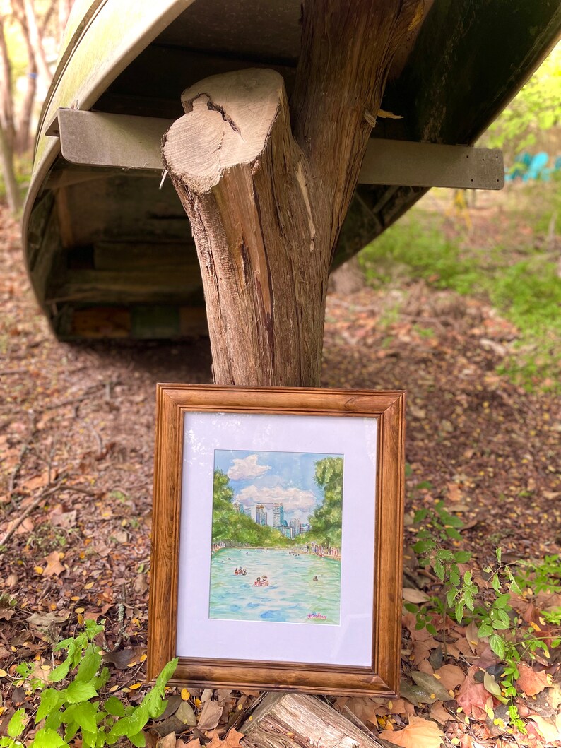K&ouml;nnte beinhalten: Ein gerahmtes Aquarellbild, das ein leuchtend blaues Schwimmbecken zeigt, umgeben von &uuml;ppigen gr&uuml;nen B&auml;umen und einer fernen Stadtsilhouette unter einem bew&ouml;lkten Himmel. Das Kunstwerk befindet sich in einem Holzrahmen mit wei&szlig;em Passepartout.