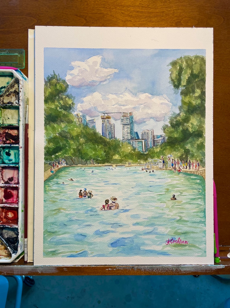 K&ouml;nnte beinhalten: Ein Aquarellgem&auml;lde, das ein &ouml;ffentliches Schwimmbad voller Menschen zeigt, umgeben von &uuml;ppigen gr&uuml;nen B&auml;umen und einer fernen Skyline unter einem blauen Himmel mit wei&szlig;en Wolken. Die Signatur der K&uuml;nstlerin, A. Cochran, ist unten rechts zu sehen.