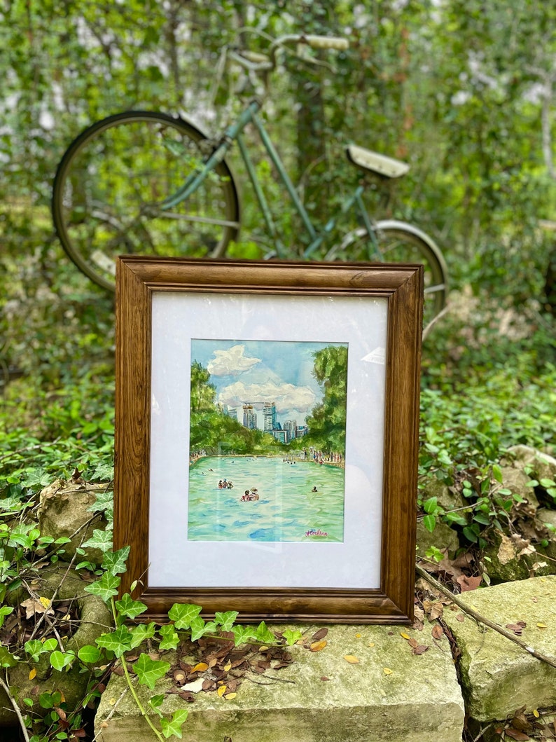 K&ouml;nnte beinhalten: Ein gerahmtes Aquarellbild, das ein t&uuml;rkisfarbenes Schwimmbecken voller Menschen zeigt, umgeben von &uuml;ppigen gr&uuml;nen B&auml;umen und einer fernen Stadtsilhouette unter einem blauen Himmel mit wei&szlig;en Wolken. Das Kunstwerk befindet sich in einem dunkelbraunen Holzrahmen.