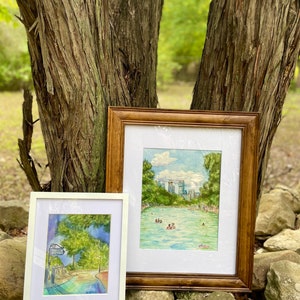 K&ouml;nnte beinhalten: Zwei gerahmte Aquarellbilder. Das kleinere Bild in einem wei&szlig;en Rahmen zeigt einen Weg mit einem Schild mit der Aufschrift Barton Springs. Das gr&ouml;&szlig;ere Bild in einem Holzrahmen zeigt ein blaues Schwimmbecken mit Menschen, B&auml;umen und einer Skyline.