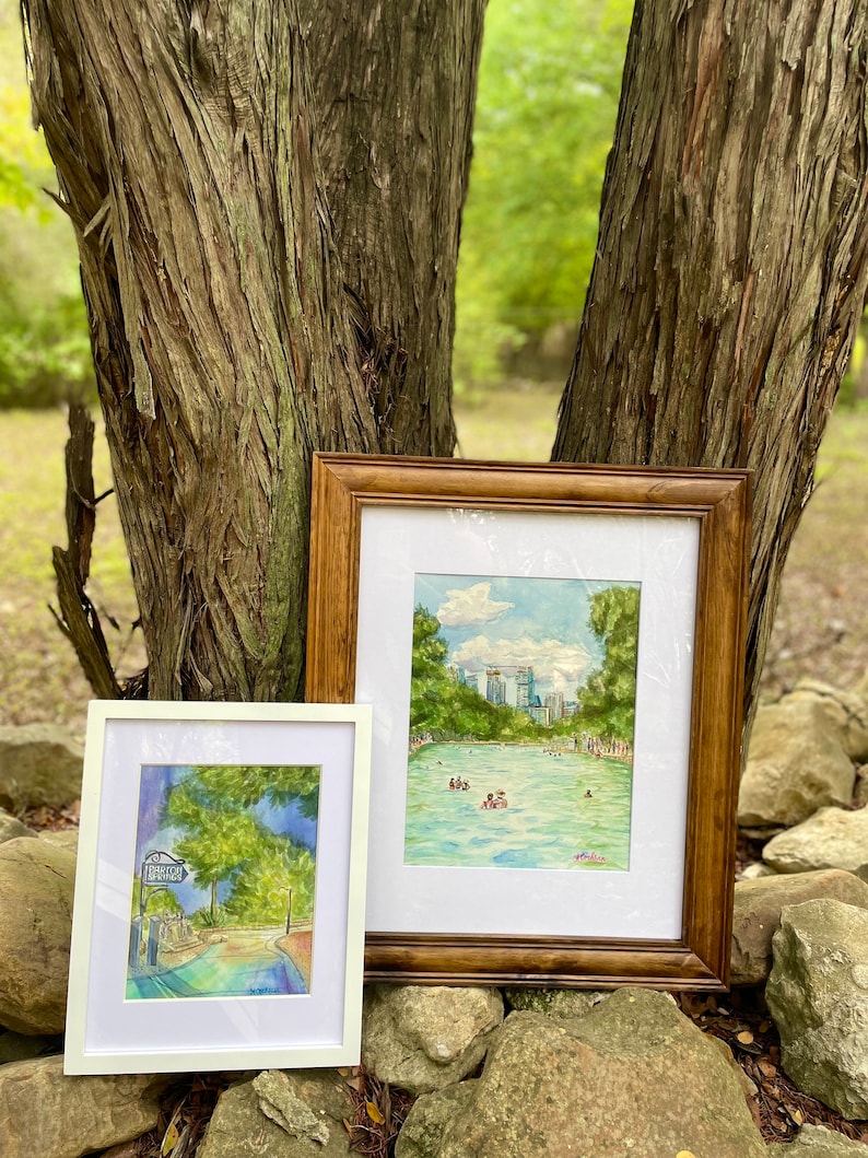K&ouml;nnte beinhalten: Zwei gerahmte Aquarellbilder. Das kleinere Bild in einem wei&szlig;en Rahmen zeigt einen Weg mit einem Schild mit der Aufschrift Barton Springs. Das gr&ouml;&szlig;ere Bild in einem Holzrahmen zeigt ein blaues Schwimmbecken mit Menschen, B&auml;umen und einer Skyline.