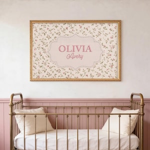 May include: A metal cot with a brass finish, white bedding, and a ruffled skirt with lace trim. Two white pillows sit inside. Above the cot hangs a rectangular framed print featuring a floral pattern and the text OLIVIA Avery.
