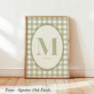 May include: A framed print featuring a light green and white gingham pattern background. An oval in the center displays a large green letter "M" with the text "is for Myles" below it. The frame is a light oak finish.