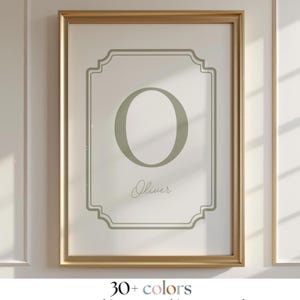 May include: A framed print with a sage green letter 'O' in a decorative frame. The name 'Oliver' is written below in a script font. The frame is gold-coloured, and the background is white. The image also includes the text '30+ colours'.