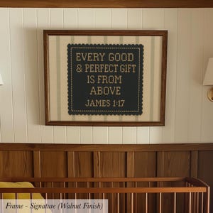 a framed cross-stitch design with the text "Every good & perfect gift is from above JAMES 1:17" on it, hanging on a wall above a wooden crib.