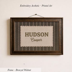 May include: A rectangular wall art piece in a bronzed walnut frame featuring a dark brown background with vertical stripes. Centered is a beige scalloped-edge rectangle with the text HUDSON Cooper in dark brown, mimicking an embroidery aesthetic.