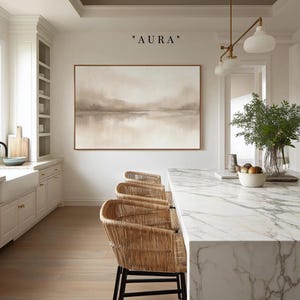 May include: A large rectangular wall art piece titled AURA featuring a soft beige and white abstract landscape painting in a thin copper frame, displayed on a white wall above a kitchen island with three woven rattan bar stools with black legs.