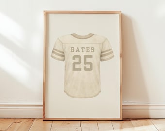 Puede incluir: Una lámina enmarcada que muestra una camiseta de fútbol americano de color beige con el nombre BATES y el número 25 impresos en color tostado oscuro. La camiseta tiene mangas cortas con dos rayas horizontales en cada brazo. Marco de roble claro.