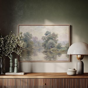 Puede incluir: Una pintura de paisaje enmarcada representa una serena escena fluvial con árboles y reflejos. La obra de arte se exhibe sobre un mueble de madera con líneas verticales decorativas. Una lámpara blanca con base dorada y un jarrón con vegetación están sobre el mueble.